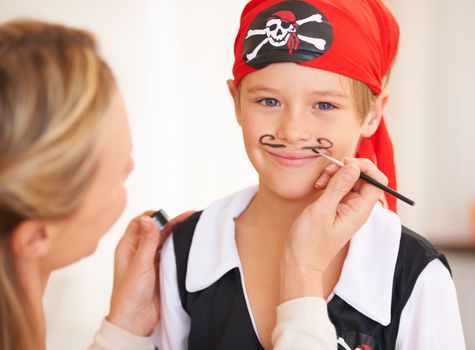 Childhood ambitions - being a pirate. Portrait of a smiling boy standing infront of his mother who is painting a mustache on his face