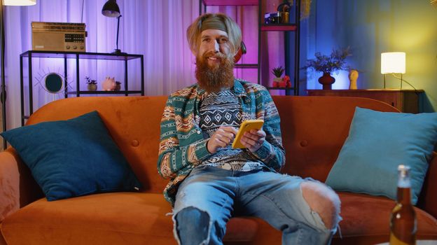 Bearded hippie man sitting on sofa uses mobile phone smiles at night home apartment. Young guy texting share messages content on smartphone social media applications online, watching relax movie