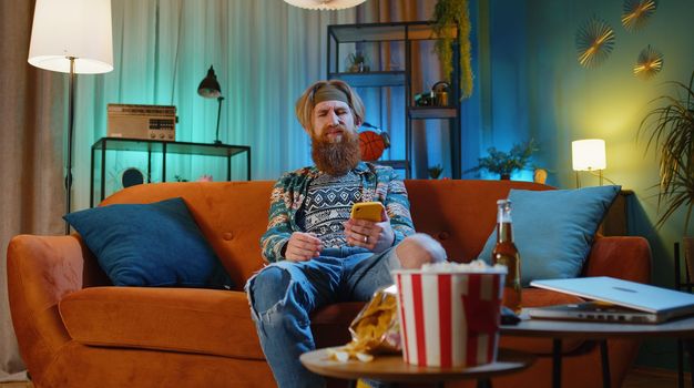 Sad bearded redhead hippie man use smartphone typing browsing, loses becoming surprised sudden lottery results, bad news, fortune loss, fail, deadline. Young ginger guy at evening home room on couch