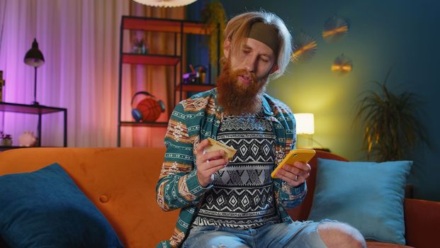 Bearded hippie man using credit bank card and smartphone while transferring money, purchases online shopping, order food delivery at home apartment indoors. Young redhead guy in evening room on couch