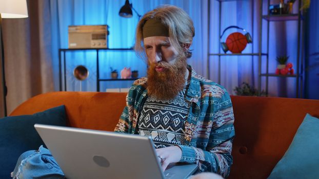 Handsome hippie bearded man freelancer at home night room sitting on couch, opens laptop start working. Young guy works on notebook, sends messages, makes online purchases, watching movies, working