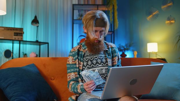 Planning budget. Rich happy hippie man counting money cash use laptop calculator, calculate domestic bills at home. Young guy satisfied of income earnings, win, saves money for planned vacation, gifts