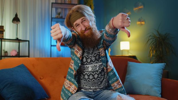 Dislike. Upset man showing thumbs down sign gesture, expressing discontent, disapproval, dissatisfied bad work at modern home apartment indoors. Displeased hippie guy in living room sitting on sofa