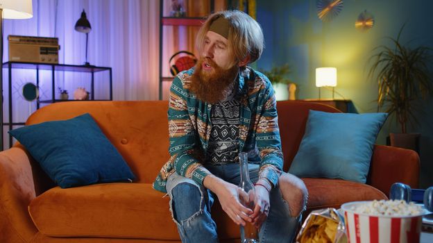 Drunk man after party. Bearded hippie redhead man at home drinks vodka beverage from bottle, problem, break up, depressed feeling bad annoyed burnout, unrequited love. Alcohol dependence addiction