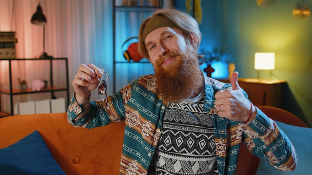 Bearded hippie redhead man real estate agent lifting hand showing the keys of new home house apartment, buying or renting property, mortgage loan at modern home apartment indoors. Guy in evening room