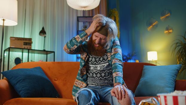 Displeased hippie redhead man at home couch rubbing temples to cure headache problem, suffering from tension, migraine, stress, grimacing in pain, high blood pressure. Hipster guy in evening room