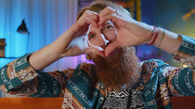 I love you. Happy bearded hippie redhead man at home couch makes symbol of love, showing heart sign to camera, express romantic feelings express sincere positive feelings. Charity, gratitude, donation
