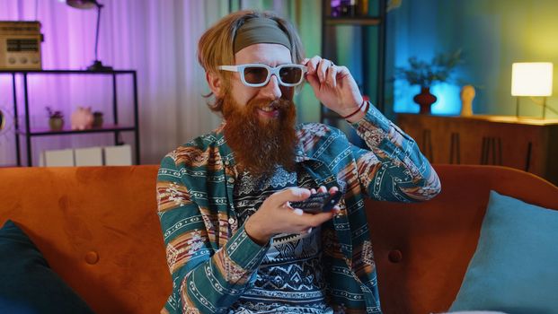 Excited bearded hippie young man sits on sofa use TV remote control watching serial, sport game, film, online social media movie content at home. Guy enjoying domestic entertainment, eating snacks