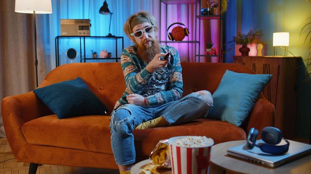Excited bearded hippie young man sits on sofa use TV remote control watching serial, sport game, film, online social media movie content at home. Guy enjoying domestic entertainment, eating popcorn