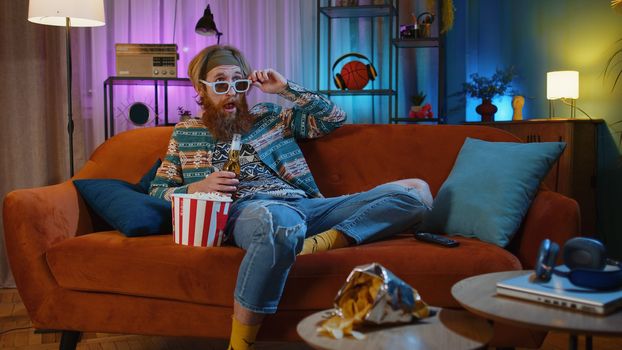 Excited bearded hippie young man sits on sofa eating popcorn snacks and watching interesting tv serial, sport game, film, online social media movie content at home. Guy enjoying domestic entertainment