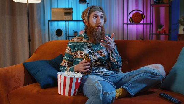 Excited bearded hippie young man sits on sofa eating popcorn snacks and watching interesting TV serial, sport game, film, online social media movie content at home. Guy enjoying evening entertainment