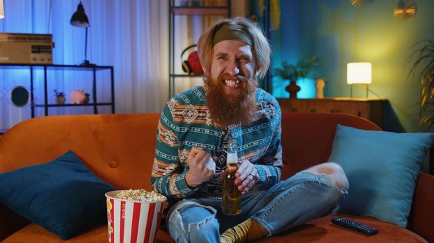 Excited bearded hippie young man sits on sofa eating popcorn snacks and watching interesting TV serial, sport game, film, online social media movie content at home. Guy enjoying evening entertainment