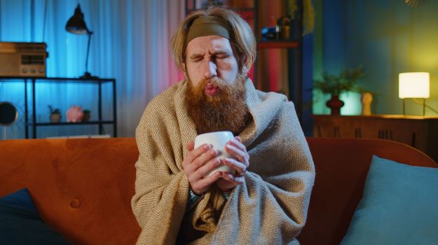 Sick man wrapped in plaid sit alone shivering from cold on sofa drinking hot tea in unheated apartment without heating due debt. Unhealthy young guy feeling discomfort try to warm up at evening home