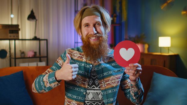 Like. Happy hippie young man looking approvingly at camera showing thumbs up, red like heart banner logo sign positive something, good great news, positive feedback. Guy on couch at home evening room