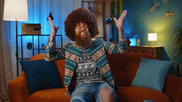 Happy bearded redhead man shouting, celebrating success, winning, goal achievement, good news raising fists in gesture I did it, lottery luck at home apartment. Young hippie guy in night room on couch