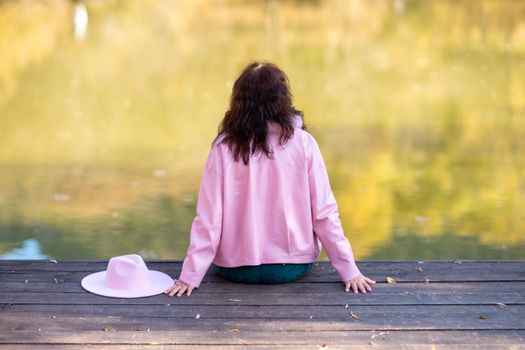 Autumn lake woman. In autumn, she sits by the pond on a wooden pier and admires nature. Dressed in a pink jacket. The concept of tourism, a weekend out of town