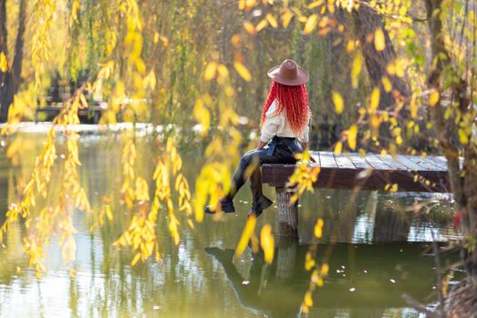 Autumn lake woman. In autumn, she sits by the pond on a wooden pier and admires nature. She is dressed in a white sweater and black leather trousers, pink curly hair and a brown hat. The concept of tourism, a weekend out of town