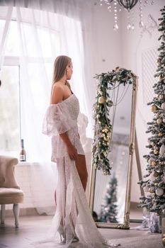 The blonde in the Christmas room. A beautiful blonde woman in a shiny light short dress with a train stands in a beautiful bright room decorated with a festive interior with a Christmas tree