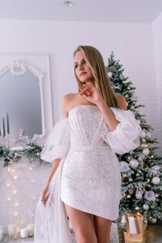 The blonde in the Christmas room. A beautiful blonde woman in a shiny light short dress with a train stands in a beautiful bright room decorated with a festive interior with a Christmas tree