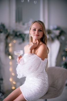 The blonde in the Christmas room. A beautiful blonde woman in a shiny light short dress with a train stands in a beautiful bright room decorated with a festive interior with a Christmas tree