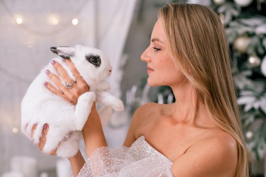 Woman holding a white rabbit symbol of the year 2023. Close-up of a beautiful young blonde woman holding a rabbit in a sparkly dress. She sits in a Christmas decorated room.