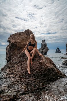 Woman swimsuit sea. Attractive blonde woman in a black swimsuit enjoying the sea air on the seashore around the rocks. Travel and vacation concept