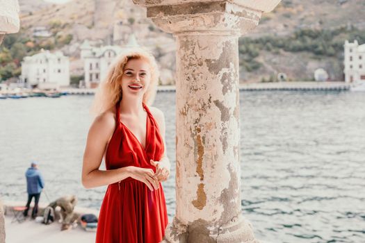 Close up shot of beautiful young caucasian woman with curly blond hair and freckles looking at camera and smiling. Cute woman portrait in a pink long dress posing on a volcanic rock high above the sea