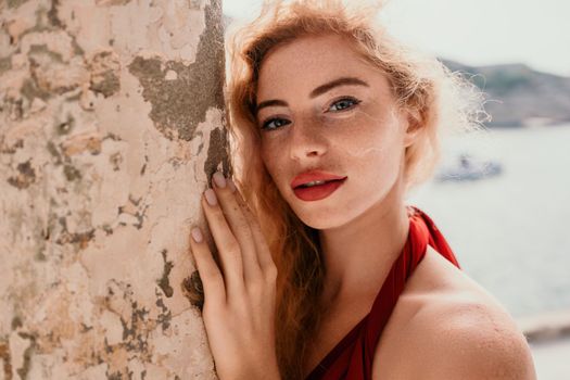 Close up shot of beautiful young caucasian woman with curly blond hair and freckles looking at camera and smiling. Cute woman portrait in a pink long dress posing on a volcanic rock high above the sea