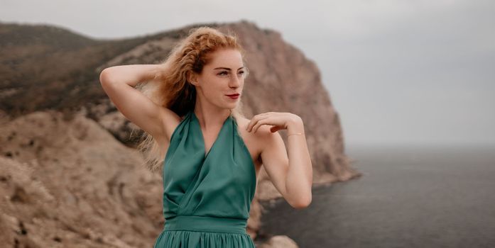 Side view a Young beautiful sensual woman in a mint long dress posing on a volcanic rock high above the sea during sunset. Girl on the nature on overcast sky background. Fashion photo