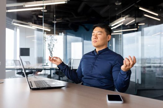 Tired young man Asian businessman meditating at work. He sits at the table in the office, took a break, closed his eyes, hands in the lotus position, relaxes.