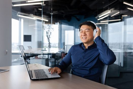 Young man Asian businessman, freelancer sits in the office at the table, works on a laptop, listens to music in white headphones, closed his eyes, enjoys, rests.