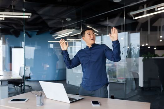 Young Asian businessman man, employee dancing while standing in office near table with laptop and documents. Raised his hands up. Celebrates victory, successful deal, monetary reward, promotion.