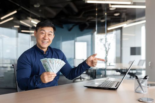 Happy young man Asian businessman sitting in the office holding cash money in his hands. He works at a laptop, points his finger at the monitor, has online meeting with partners, investors, smiles.