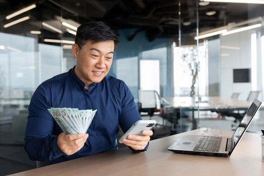 A young Asian businessman is sitting in the office at a table with a laptop. He is holding a phone and cash money. Plays online games, checks account, successful deal, investment.