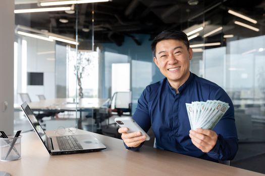 A young Asian man is sitting in the office. He is holding a phone and cash money. He looks at the camera, smiles.