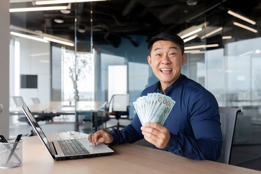 A young Asian businessman is sitting in the office at a table with a laptop. Holds cash money in hands. Plays online games, places bets, checks account, successful deal, investment. He looks at the camera, smiles.