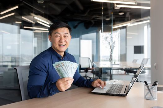 Happy young man Asian businessman sitting in the office at the table holding cash money in his hands, showing it to the camera, smiling. Works on a laptop and documents.