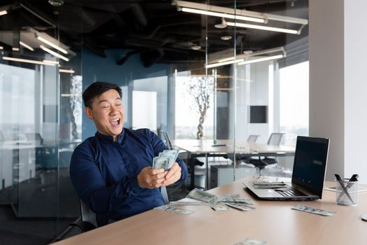 A happy young Asian man, an office worker, an entrepreneur holds cash money in his hands, received a salary, a bonus. He is sitting at a table in the office, rejoices, shouts with joy.