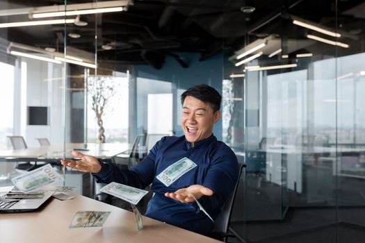 A happy young Asian man sits in the office at the table and throws cash money, dollars in the air. Celebrates winnings, earnings, wealth.