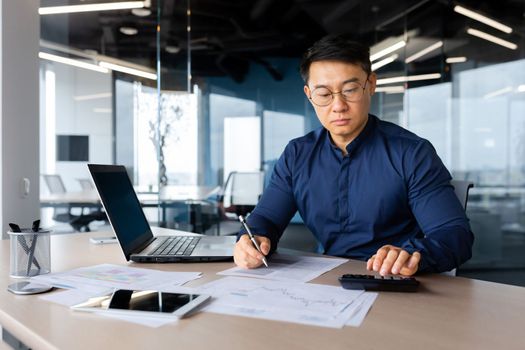 Portrait of a serious young Asian accountant, banker. Sits working in the office with documents, laptop, calculator.