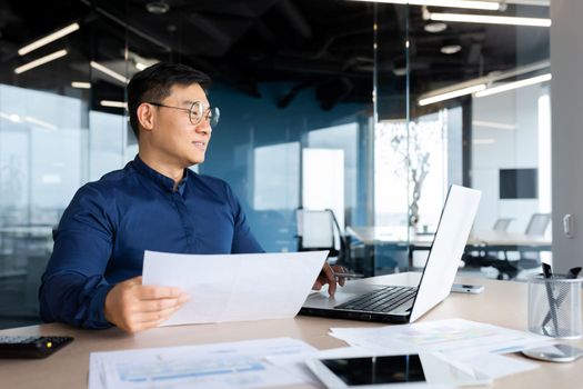 A young Asian journalist, blogger, freelancer is sitting in the office at the table. Works on a laptop and with papers. Writes an article, a book, an interview.