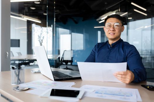 Portrait of a young Asian architect, designer, engineer. Sitting at the desk in the office, holding papers. Works with documents, projects, plans. He looks at the camera, smiles.