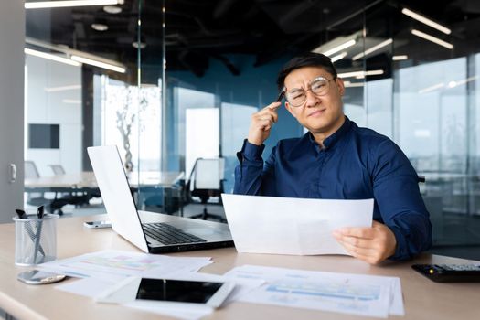 Serious young Asian architect, designer, engineer. Sitting at the desk in the office, holding documents and a pencil. The problem is working with projects, plans. Looking worriedly at the camera.