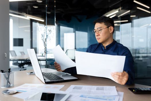 A serious young man, an Asian businessman, works in the office at the table, holds and looks at documents, graphs, plans, bills.