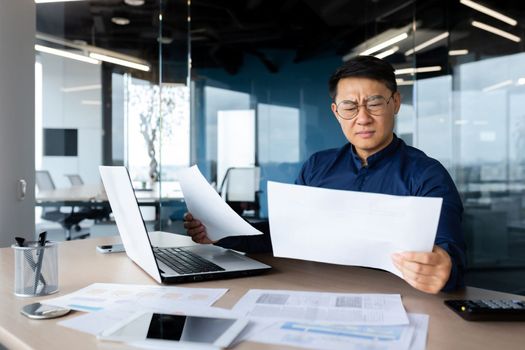 Problems at work. Portrait of a worried young Asian man accountant, banker. He sits and works in the office, holding documents, bills, loans in his hands. Works with a laptop, calculator.