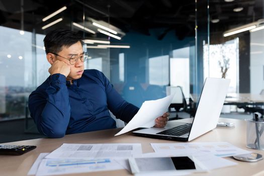 Tired young Asian man, accountant, businessman, freelancer. He sits in the office at a table with a laptop, looks through documents, holds his head in his hands, sighs a hard and boring day.