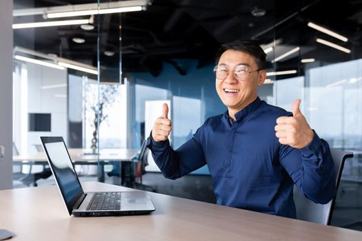 Portrait of a young handsome man, an Asian designer, engineer, freelancer. He sits in the office at the table with a laptop, looks at the camera, smiling, shows his hands with his fingers super.