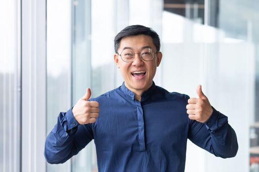 Portrait of a happy young handsome Asian man, businessman, student in glasses and a blue shirt standing in the office, looking at the camera, smiling, showing his hands super.