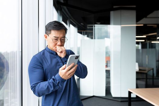 A worried and serious young Asian man is standing in the lobby of an office, hospital, bank. Holds the phone in his hands, looks at the screen in shock, received and reads bad news, dials a message.
