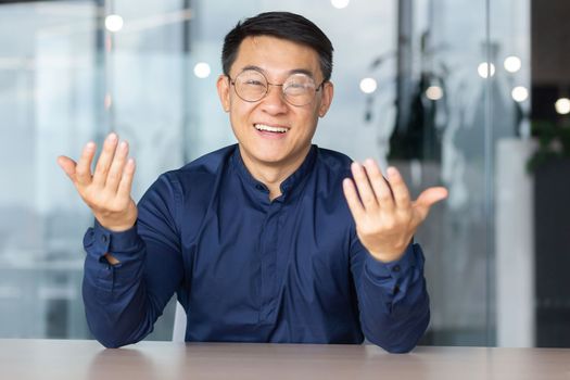 Portrait young male Asian teacher is lecturing students, taking exams online video call. Sitting in the office at the table, smiling at the camera, explains, waving his hands.